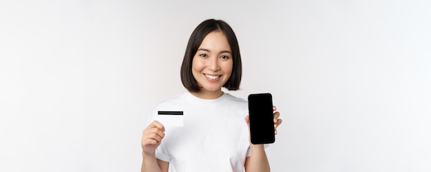 흰색 배경 위에 서 있는 휴대폰 화면과 신용 카드를 보여주는 웃고 있는 젊은 아시아 여성의 초상화