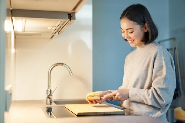 부엌에 서서 자신을 위해 요리하는 샌드위치를 만드는 웃는 젊은 아시아 여성의 초상화