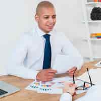 Free photo portrait of a smiling young businessman with business reports on table sitting with his coworker