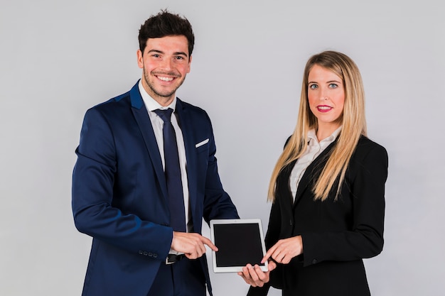 Free photo portrait of a smiling young businesswoman and businessman pointing digital tablet against grey backdrop