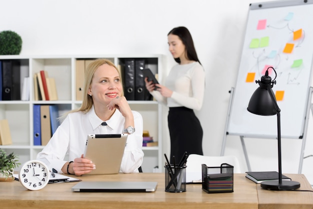 Free photo portrait of a smiling young businesswoman holding digital tablet looking away with her colleague standing at background