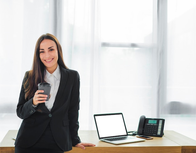 Portrait of a smiling young businesswoman standing in front of desk holding disposable coffee cup