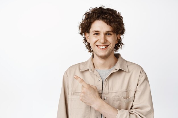 Portrait of smiling young caucasian man with curly face and white teeth pointing finger at upper left corner showing advertisement white background