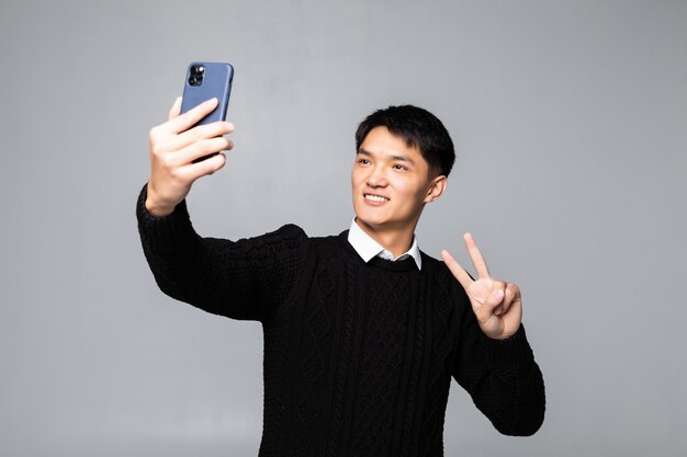 흰 벽 위에 격리하는 동안 휴대 전화로 셀카를 복용 웃는 젊은 중국 남자의 초상화