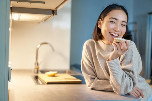 家にいる笑顔の若い幸せな女性のポートレート、キッチンに立ってトーストを食べ、