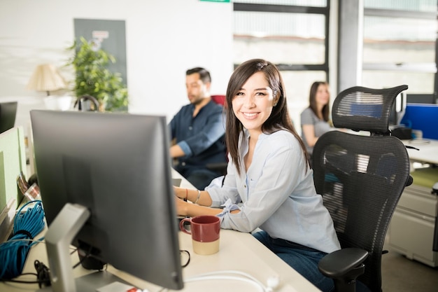 Ritratto di giovane donna latina sorridente seduta alla sua scrivania con i colleghi che lavorano sul retro