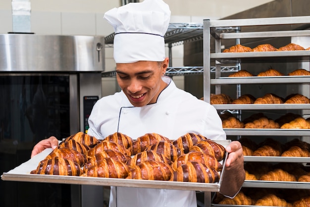 Foto gratuita ritratto di un giovane panettiere maschio sorridente guardando croissant appena sfornato in teglia