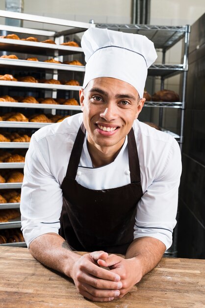 Portrait of a smiling young male baker in uniform leaning on table in the bakery