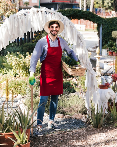 Portrait of a smiling young male gardener holding gardening tool and basket