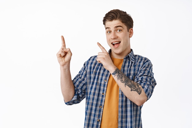 Portrait of smiling young man pointing fingers at upper left corner standing over white.