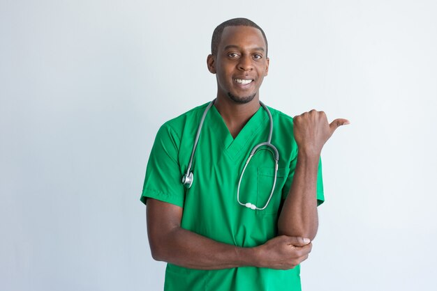 Portrait of smiling young physician pointing at something with thumb. 