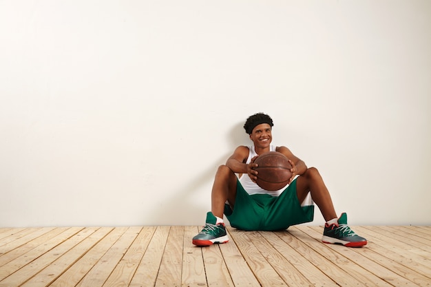 A portrait of an smiling young player sitting on the wooden floor against a white wall holding an old brown basketball