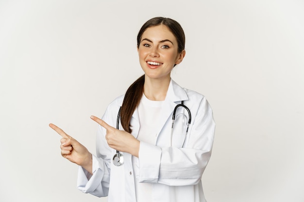 Portrait of smiling young woman doctor healthcare medical worker pointing fingers left showing clini...