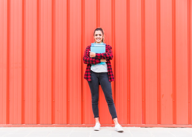 Portrait of a smiling young woman holding books in hand standing against an orange wall
