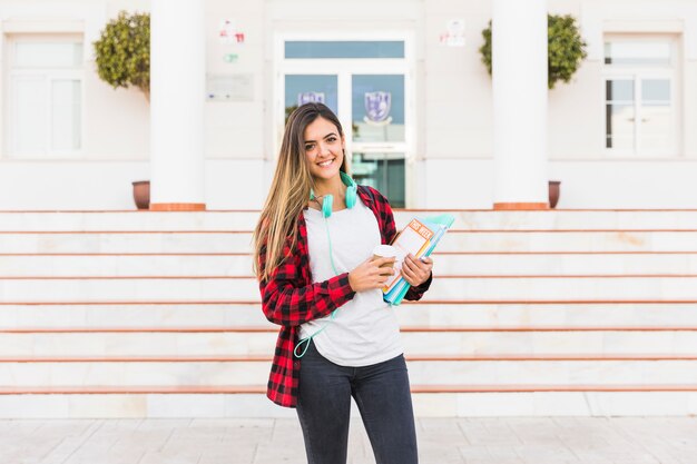 Portrait of a smiling young woman holding books in hand standing against university building