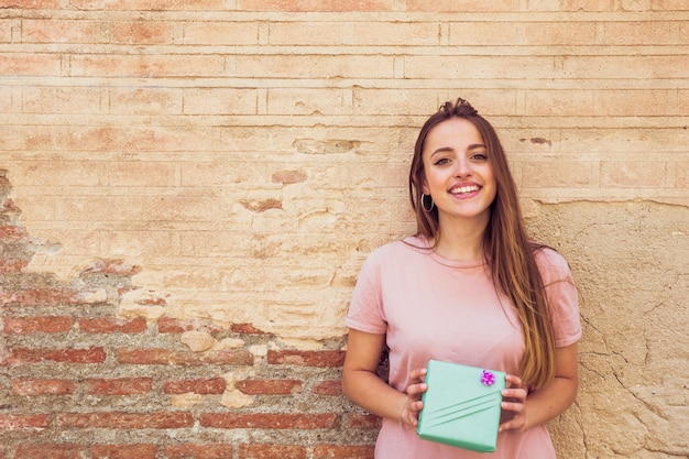 Portrait of a smiling young woman holding gift in front of old wall