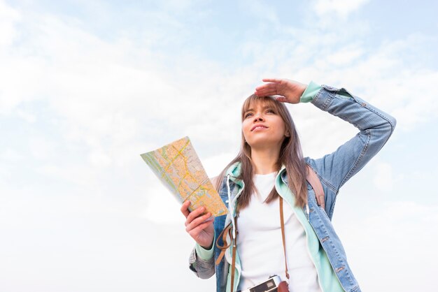 Free photo portrait of a smiling young woman holding map in hand shielding her eyes against sky