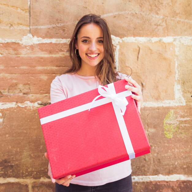 Portrait of a smiling young woman holding red gift box against wall