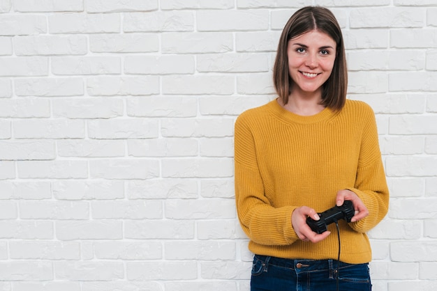 Портрет улыбающейся молодой женщины, играющей в видеоигру с джойстиком, стоящим против белой кирпичной стены