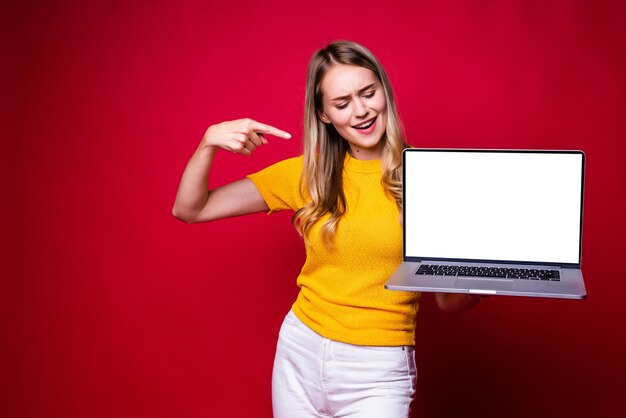 Portrait of smiling young woman pointed on screen of laptop pc computer isolated on red wall.