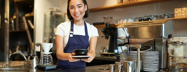 Portrait of smiling young woman providing customer service in cafe holding cup of coffee giving