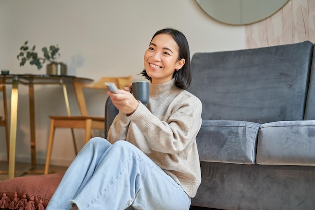 바닥에 앉아 TV를 보고 컵에서 커피를 마시는 TV 근처 집에서 쉬고 웃는 젊은 여성의 초상화