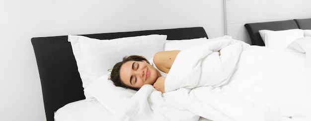 Free photo portrait of smiling young woman snugging in bed hugging her duvet lying in white linen sheets and