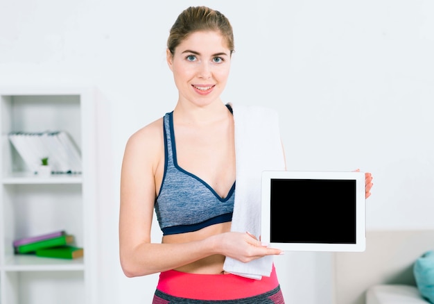 Foto gratuita ritratto della giovane donna sorridente in abiti sportivi con l'asciugamano sopra la spalla che mostra compressa digitale