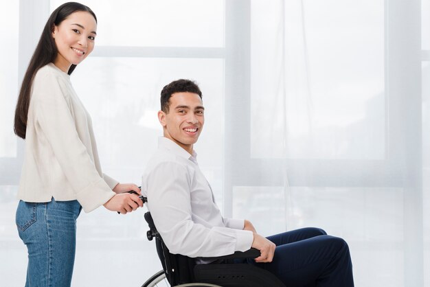 Portrait of a smiling young woman standing behind the man sitting on wheel chair looking at camera