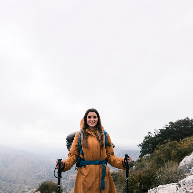 ハイキングの棒を保持している山の上に立っている笑顔の若い女性の肖像画