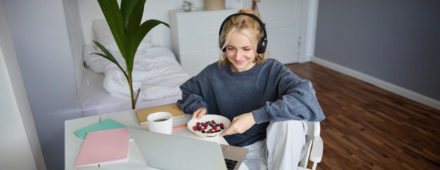 Portrait of smiling young woman watching tv show in headphones eating breakfast and looking at