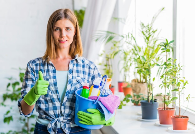 Portrait of a smiling young woman with bucket of cleaning equipments gesturing thumbs up