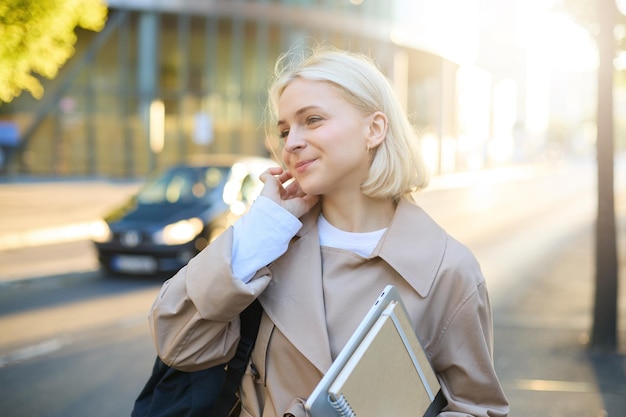 Free photo portrait of smiling young woman with notebook tucks hair behind her ear walking along street female