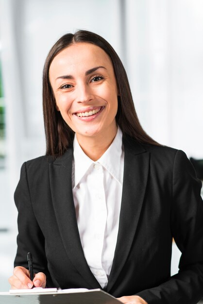 Portrait of smiling young woman writing with pen on clipboard looking at camera