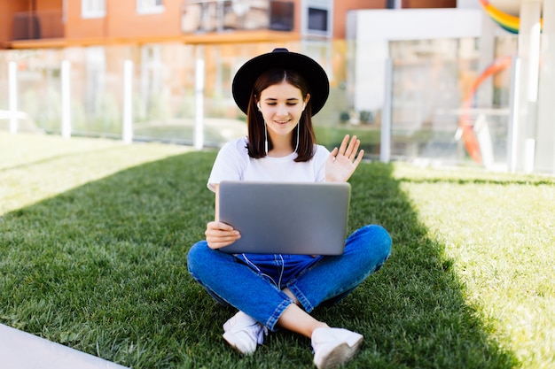 Portrait of sociable woman sitting on green grass in park with legs crossed during summer day while using laptop and wireless earpbud for video call