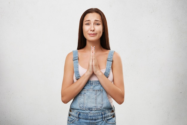 Foto gratuita ritratto di donna sorridente che indossa una camicia bianca e una tuta di jeans, tenendo le mani unite, andando a piangere chiedendo perdono. giovane donna castana con lo sguardo imbronciato isolato sopra la parete bianca