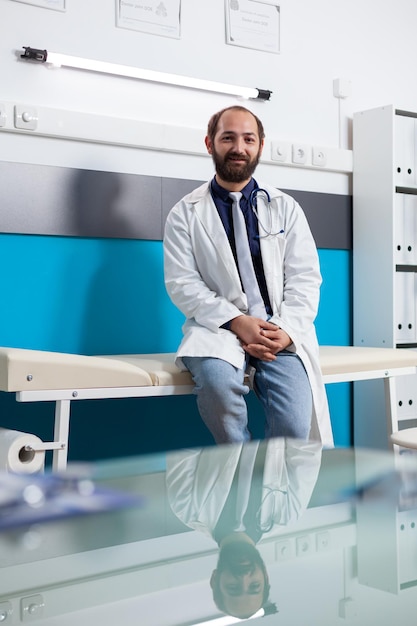 Portrait of specialist wearing uniform in medical cabinet, waiting to start health appointment with patient. Physician sitting on bed to consult people with illness, giving support and advice.