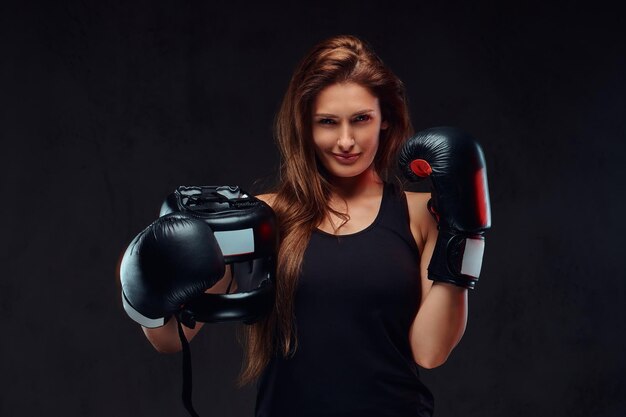 Portrait of a sportive woman dressed in sportswear wearing boxing gloves holds protective helmet. Isolated on a dark textured background.