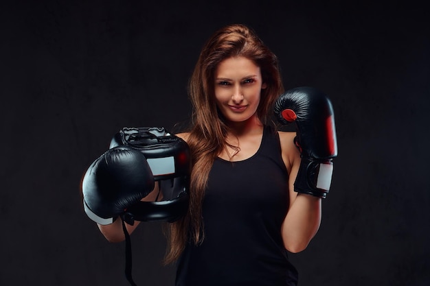 Portrait of a sportive woman dressed in sportswear wearing boxing gloves holds protective helmet. Isolated on a dark textured background.