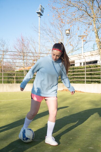 フィールドでボールを蹴るスポーティな10代の少女の肖像画。新しい試合の準備のためにパスを練習している彼女の女の子のチームとサッカーをしているスポーツウェアの真面目な女の子。健康的なライフスタイルとスポーツのコンセプト