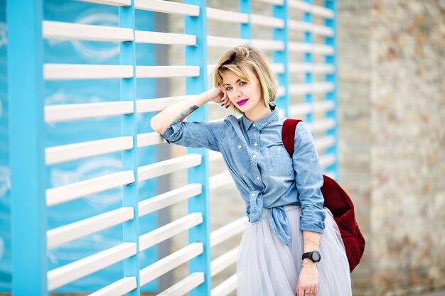 Portrait of a standing smiling woman with short blond hair, bright pink lips and nude make up leaning on blue and white stripes fence