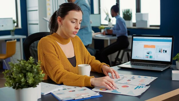 Portrait of startup employee comparing two sheets of paper with charts analyzing financial data while drinking morning coffee. Woman working in busy marketing department sitting at desk with laptop.