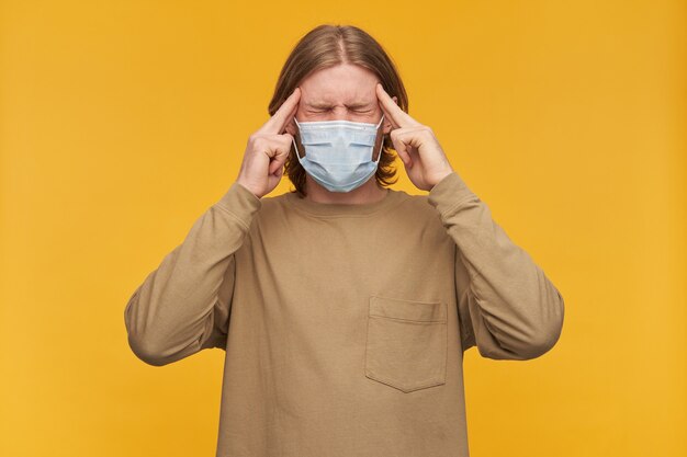 Portrait of stressed, adult male with blond hair and beard. wearing beige sweater and medical protective face mask. massaging temples, suffer from headache. stand isolated over yellow wall