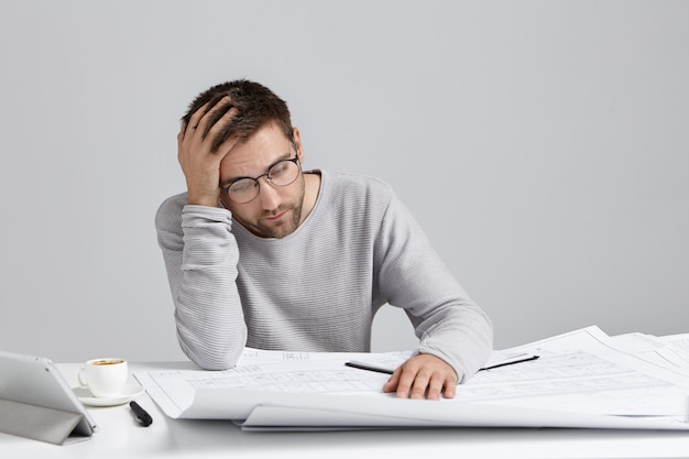 Portrait of stressed young Caucasian architect holding hand on head and looking at blueprints