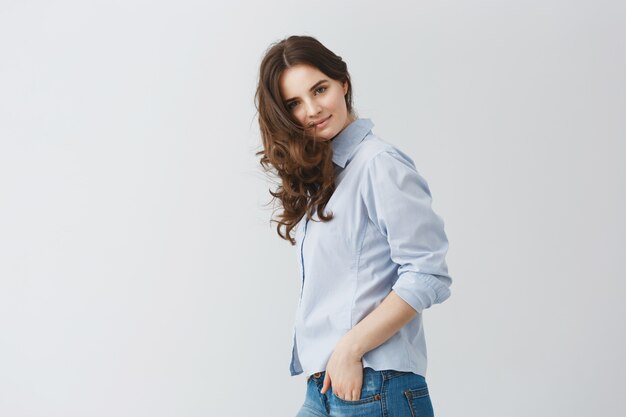 Portrait of student girl with dark long wavy hair holding hands in pockets with calm and happy face expression.