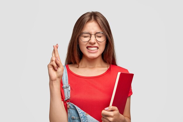 Portrait of student with long hair keeps fingers crossed for good luck on exam, holds textbook