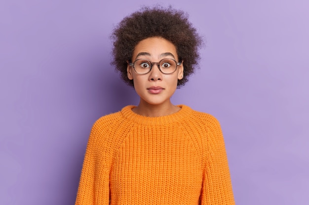 Portrait of stunned curly millennial girl stares through spectacles dressed in orange sweater reacts on shocking news.