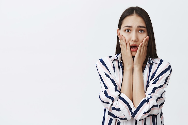 무료 Photo 스트라이프 블라우스에 기절하고 강렬한 잘 생긴 여학생의 초상화, 헐떡이며, 뺨에 손바닥을 들고, 가슴 아픈 이야기를 동정하면서 회색 벽 위에 포즈를 취함
