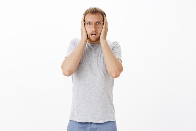 Free photo portrait of stunned and shocked intense adult male in glasses holding hands on head gasping and opening mouth from shock making terrible mistake starting panic over white wall