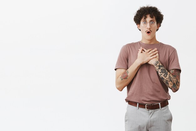 Portrait of stunned shocked stylish and artistic young jewish guy with tattooed arm moustache and curly dark hair in glasses and trendy outfit holding hands on breast gasping from shook and worry